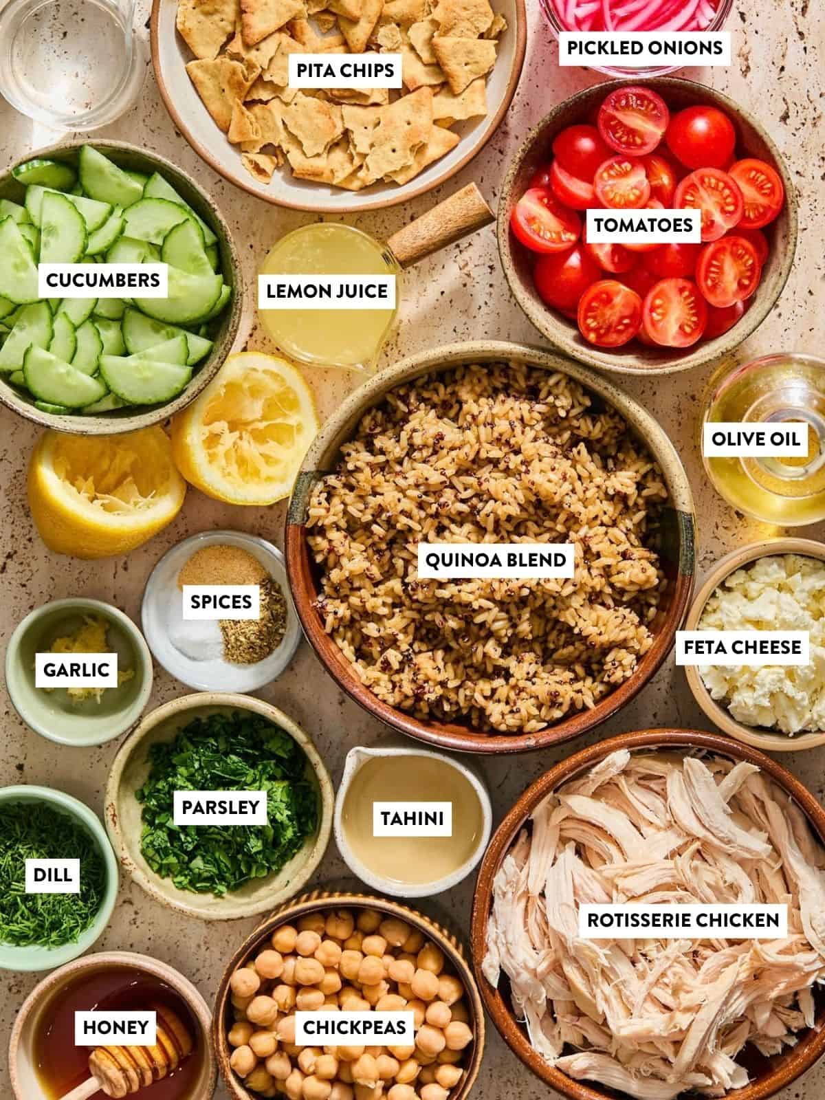 Top-down view of bowls with labeled ingredients: pita chips, pickled onions, cucumbers, lemon juice, tomatoes, olive oil, feta cheese, quinoa blend, garlic, spices, parsley, tahini, dill, rotisserie chicken, honey, and chickpeas.