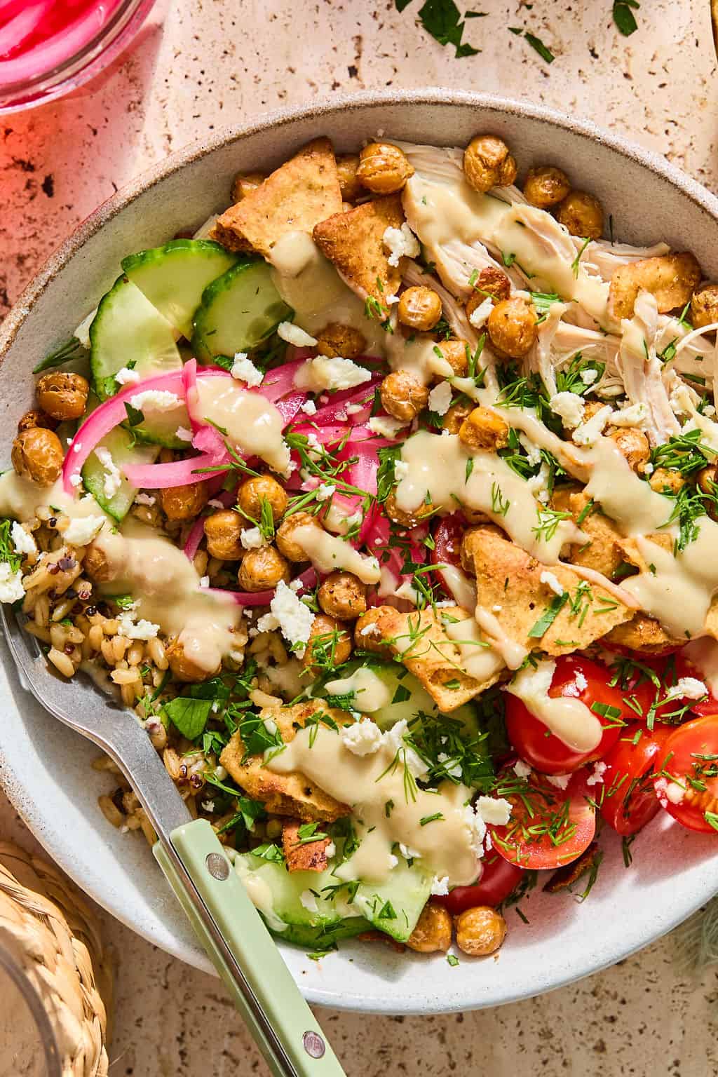 A greek chicken bowl with shredded chicken, cucumber, tomato, pickled onions, crispy chickpeas, pita chips, fresh herbs, feta cheese, and a creamy dressing, served with a fork.