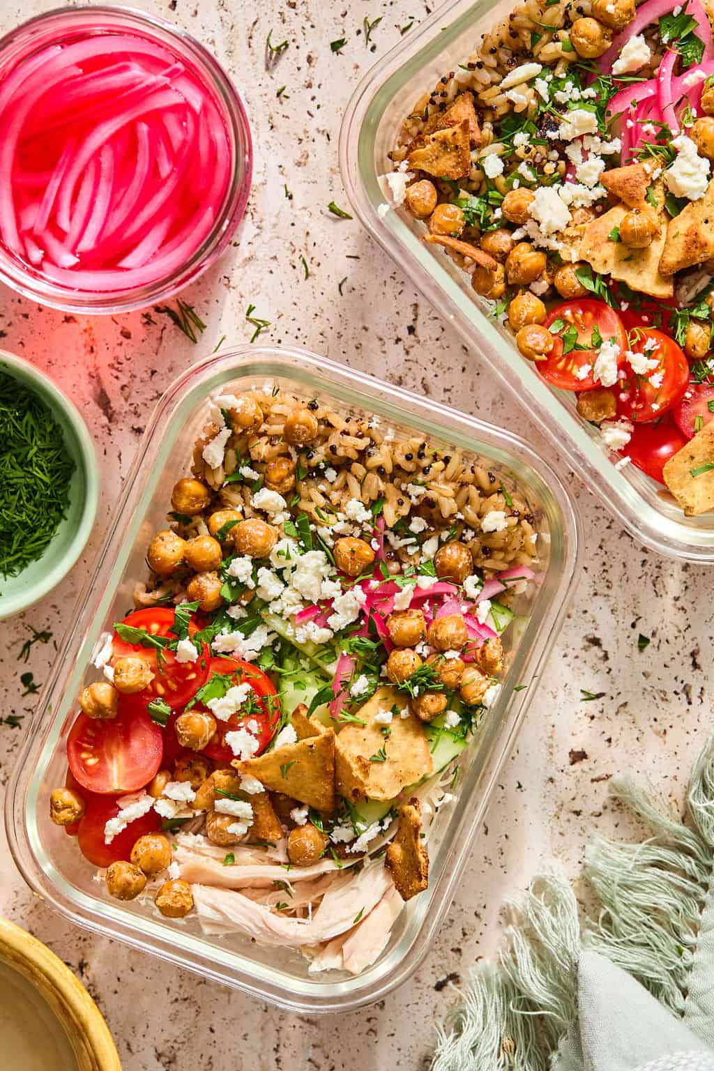 Two glass containers filled with rice, shredded chicken, cherry tomatoes, lettuce, roasted chickpeas, pita chips, pickled onions, and feta cheese creating greek chicken bowls. A jar of pickled onions and a small bowl of herbs are nearby.