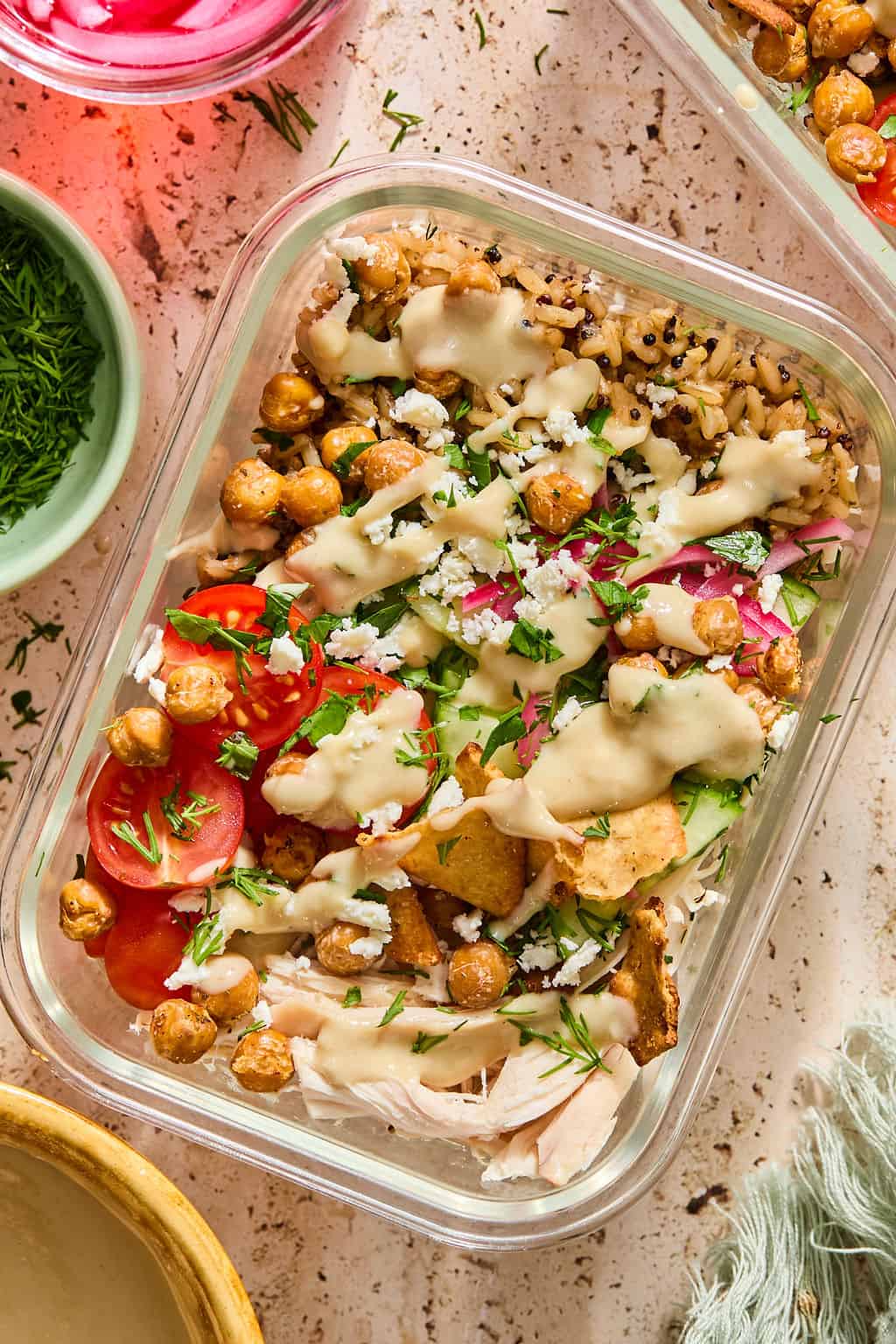 A glass container filled with a salad of cherry tomatoes, chickpeas, crumbled cheese, herbs, shredded chicken, pita chips, and grains, topped with a creamy dressing.