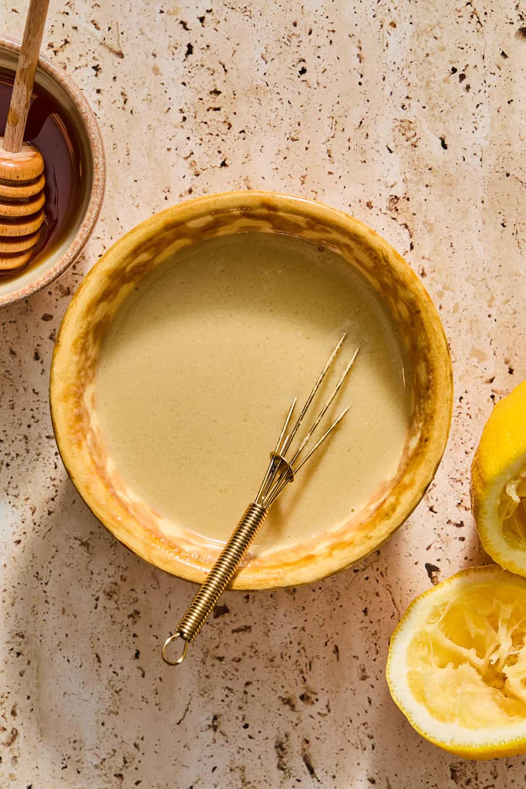 A bowl of creamy dressing with a small whisk, surrounded by a honey dipper, honey, and squeezed lemon halves on a textured beige surface.