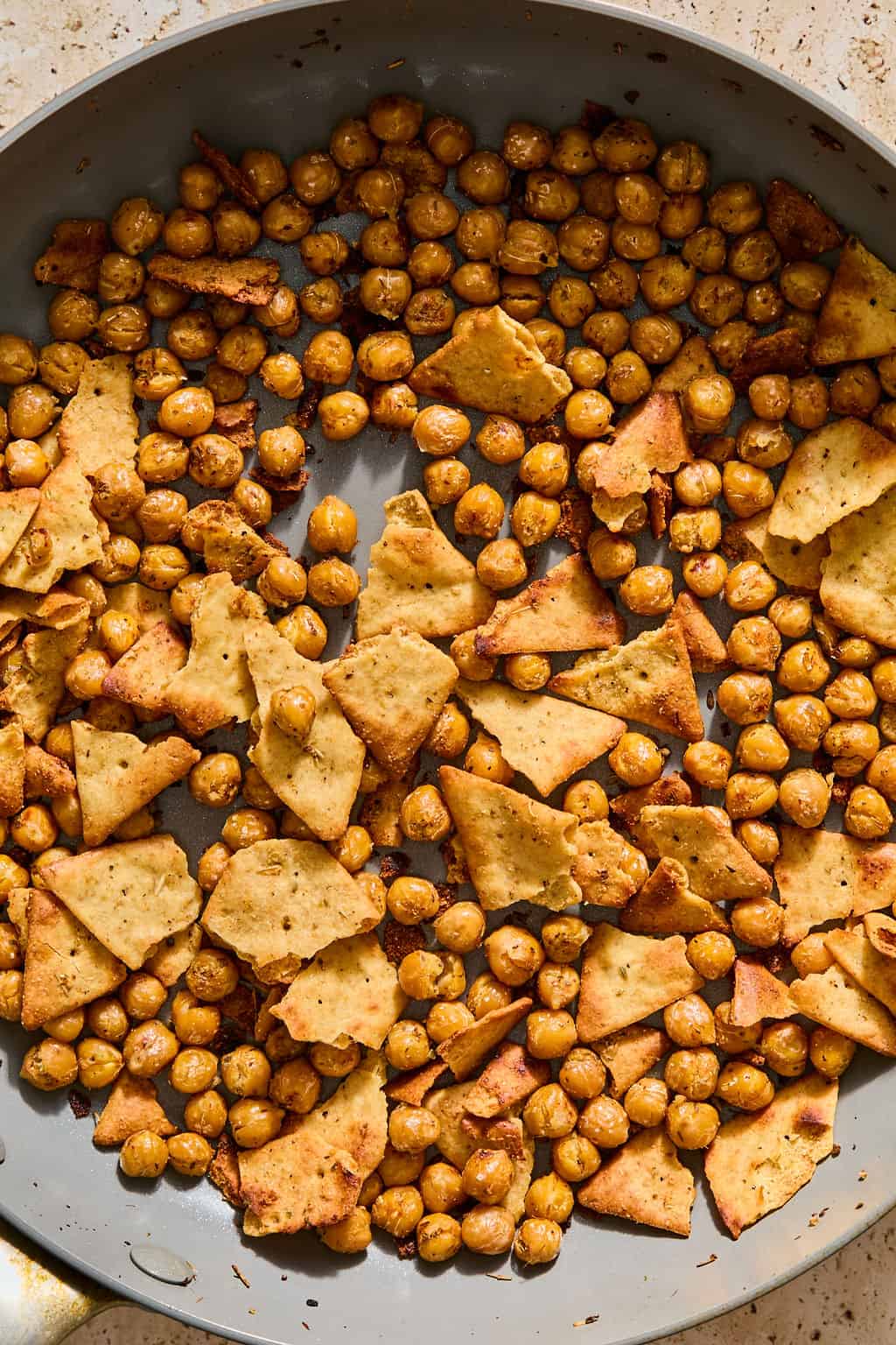 Chickpeas and broken pieces of pita chips, seasoned and roasted, are spread out evenly on a round, gray baking sheet.