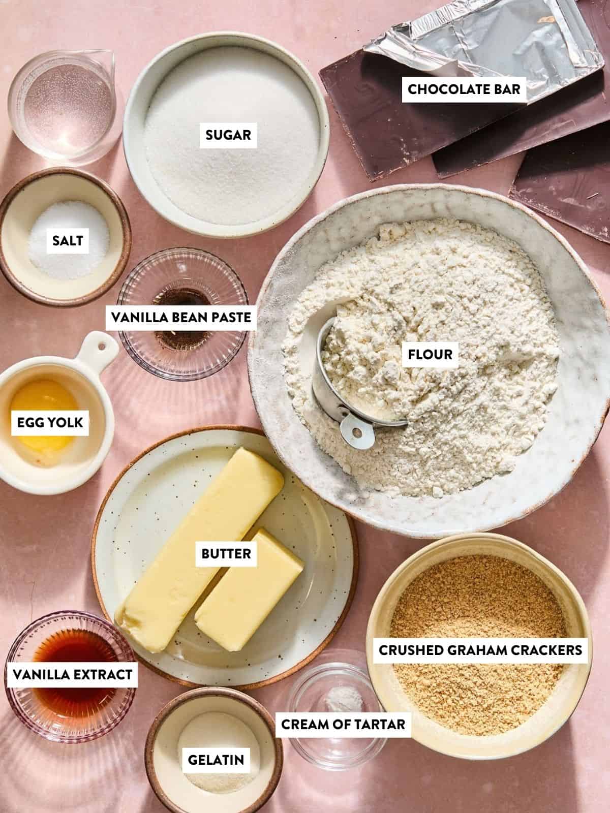 An overhead view of baking ingredients labeled: chocolate bar, sugar, salt, vanilla bean paste, egg yolk, flour, butter, vanilla extract, gelatin, cream of tartar, and crushed graham crackers.