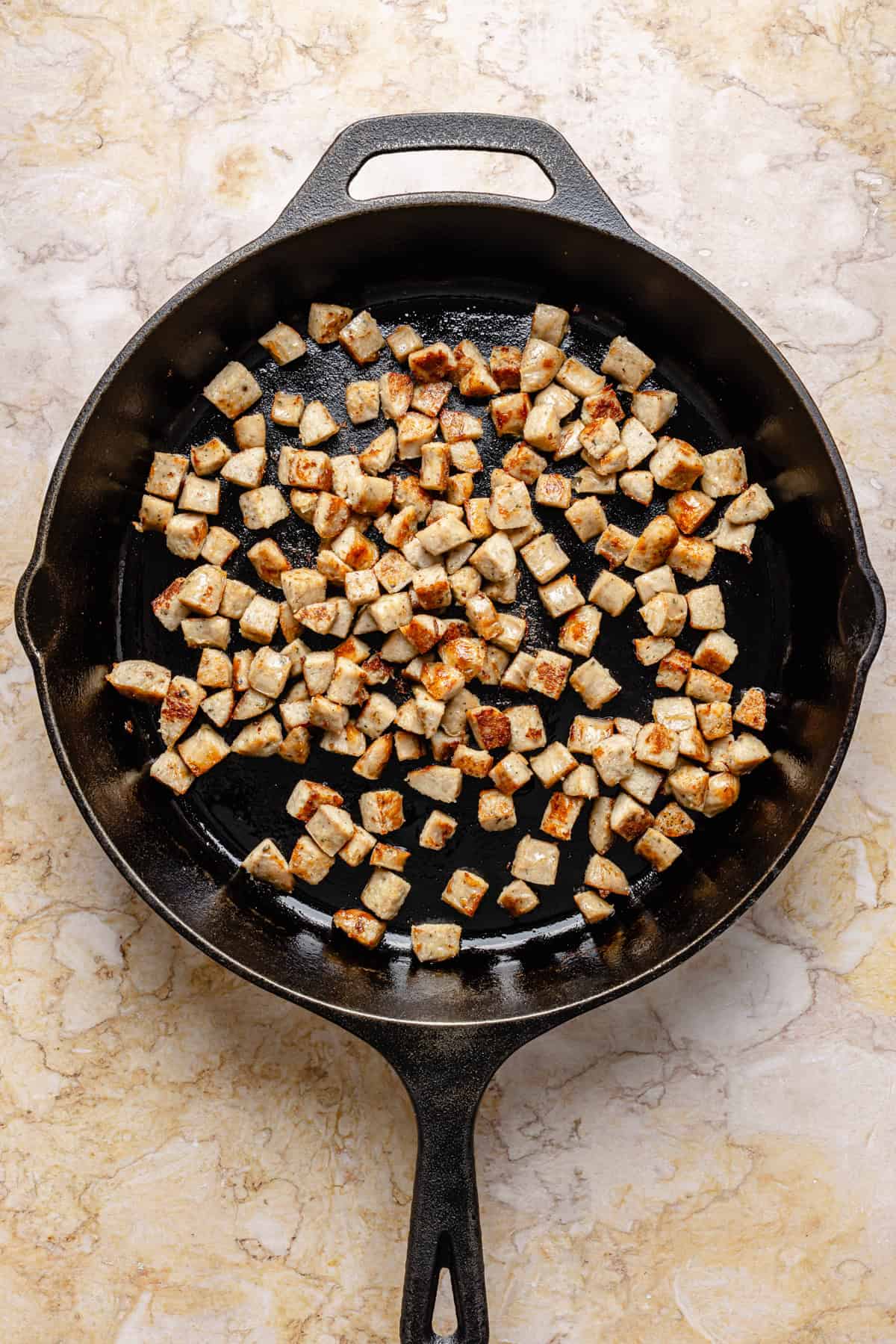 Diced sausage pieces are browning in a black cast iron skillet on a light-colored marble surface.