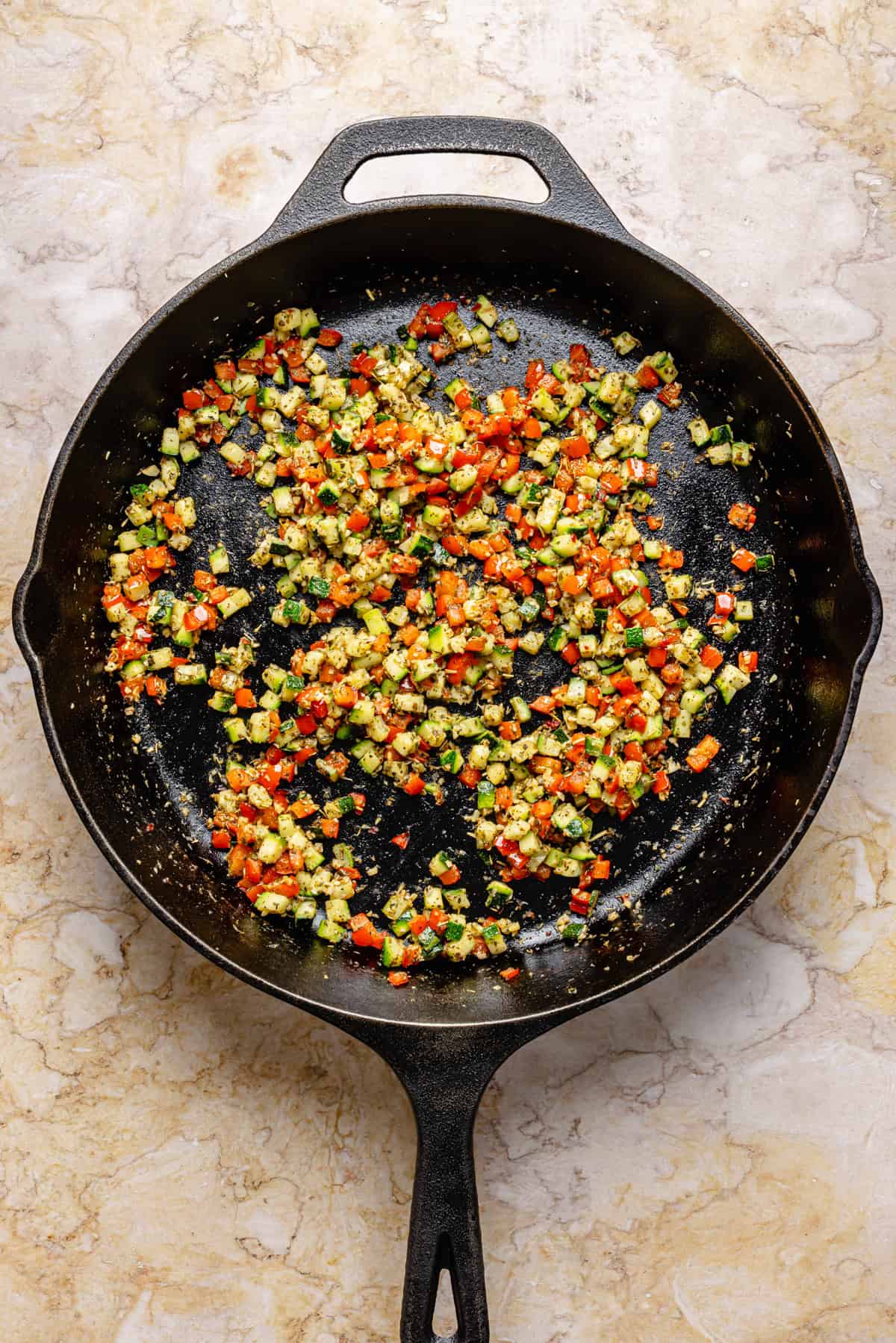 Diced vegetables, including red and green peppers, are sautéing in a black cast-iron skillet on a light marble surface.