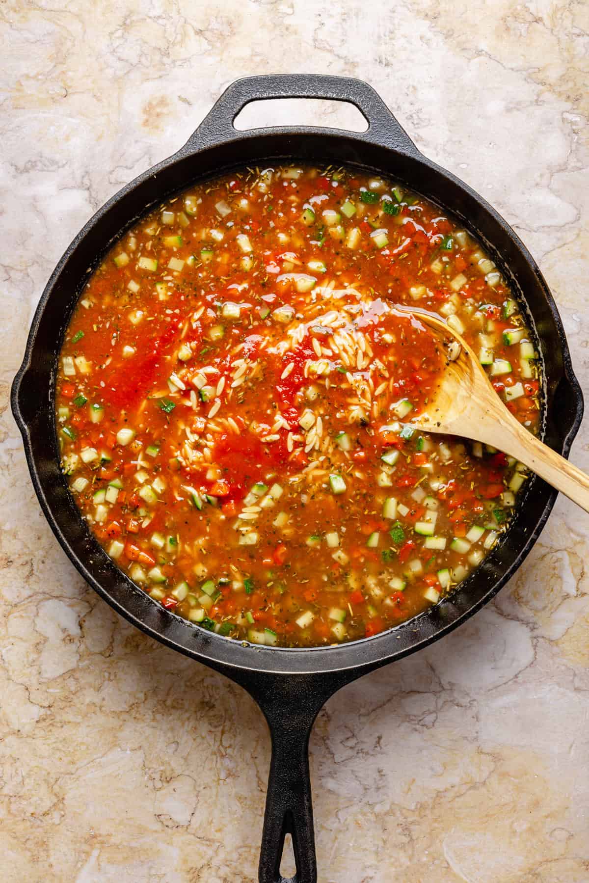A cast iron skillet filled with a colorful mixture of chopped vegetables, rice, and tomato-based broth. A wooden spoon rests inside, blending the ingredients. The skillet sits on a light, marbled surface.
