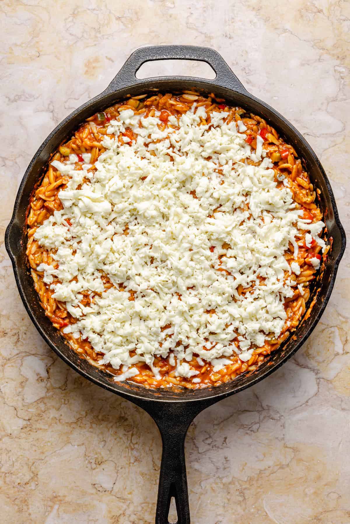 A cast iron skillet filled with cooked orzo in tomato sauce, topped with a generous layer of shredded mozzarella cheese, sits on a light-colored marble surface.
