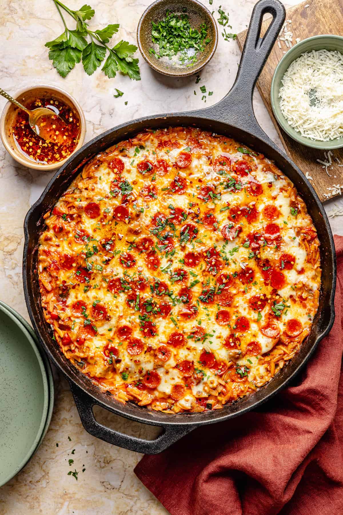 One pot pizza orzo in a cast-iron skillet, topped with melted cheese and herbs, surrounded by bowls of grated cheese, chili oil, chopped herbs, and an empty green plate with a red napkin.