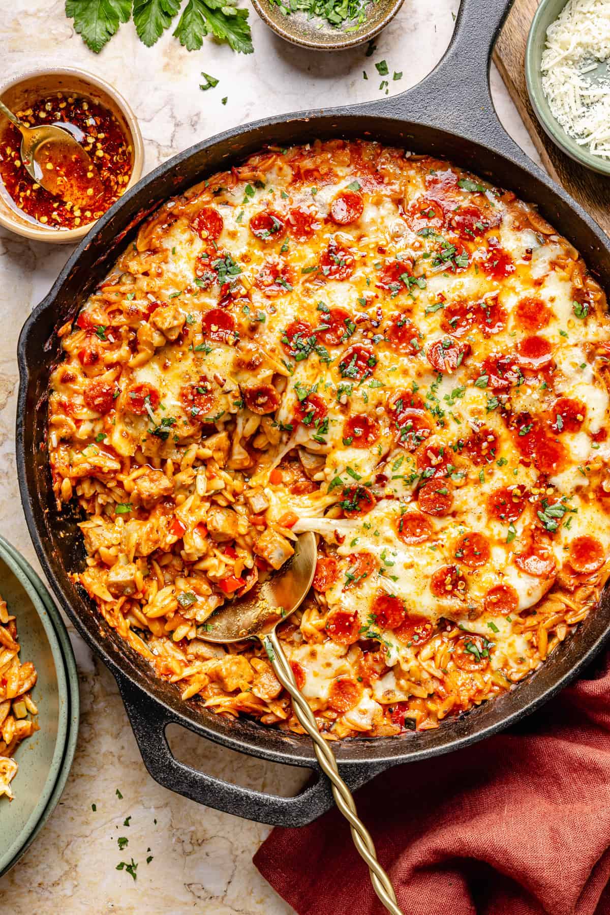 A skillet filled with one pot pizza orzo topped with mini pepperoni, melted cheese, and herbs. A serving spoon rests in the dish, with some pasta scooped out. Bowls, herbs, and sauce are nearby on the table.