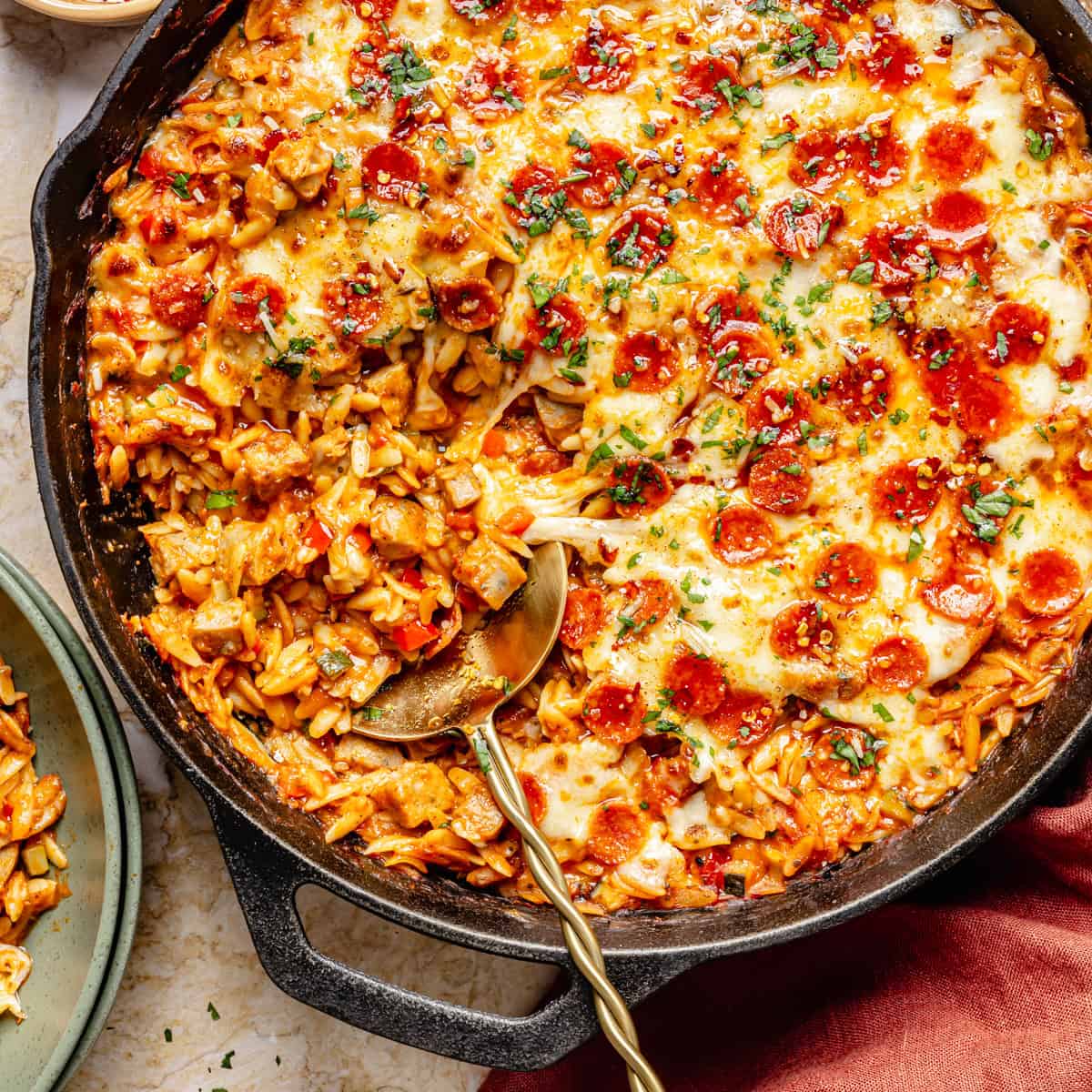 One pot skillet filled with pizza orzo topped with melted cheese and pepperoni, with a serving spoon lifting a portion onto a plate.