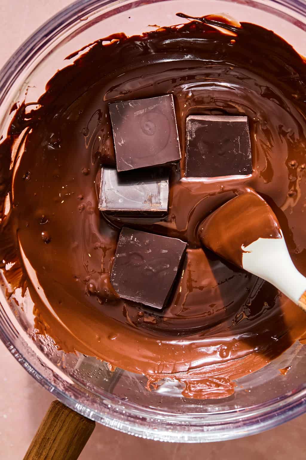 A silicone spatula stirring solid dark chocolate squares into a bowl of melted chocolate to temper it.