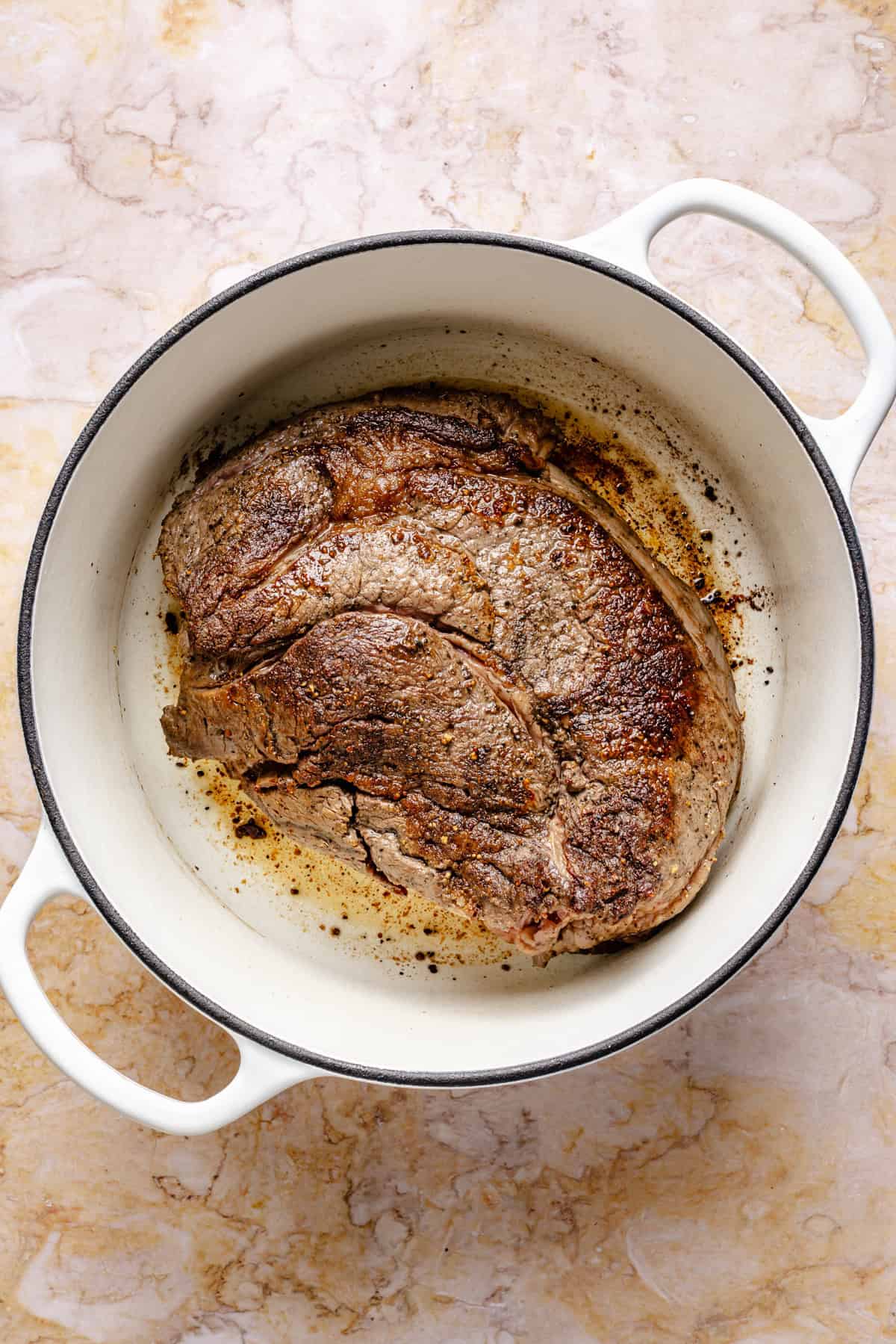 A browned beef roast sits in a white Dutch oven on a light marble surface, with seared edges visible and some browned bits around the meat.