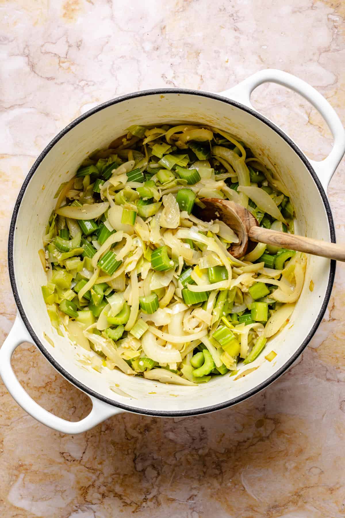 White pot filled with sautéed sliced leeks and onions, stirred with a wooden spoon, on a light marble surface.