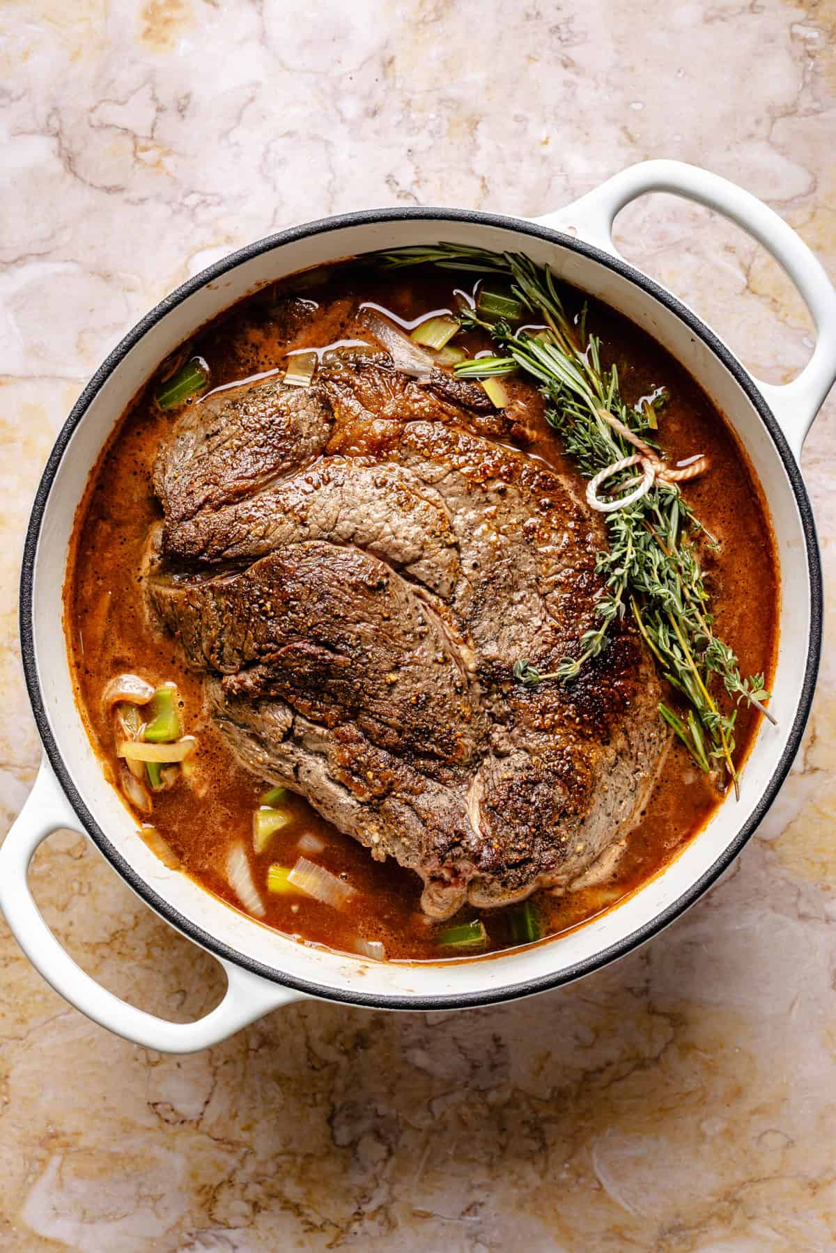 A cooked pot roast sits in a white Dutch oven with broth, herbs, and chopped vegetables on a marble surface.
