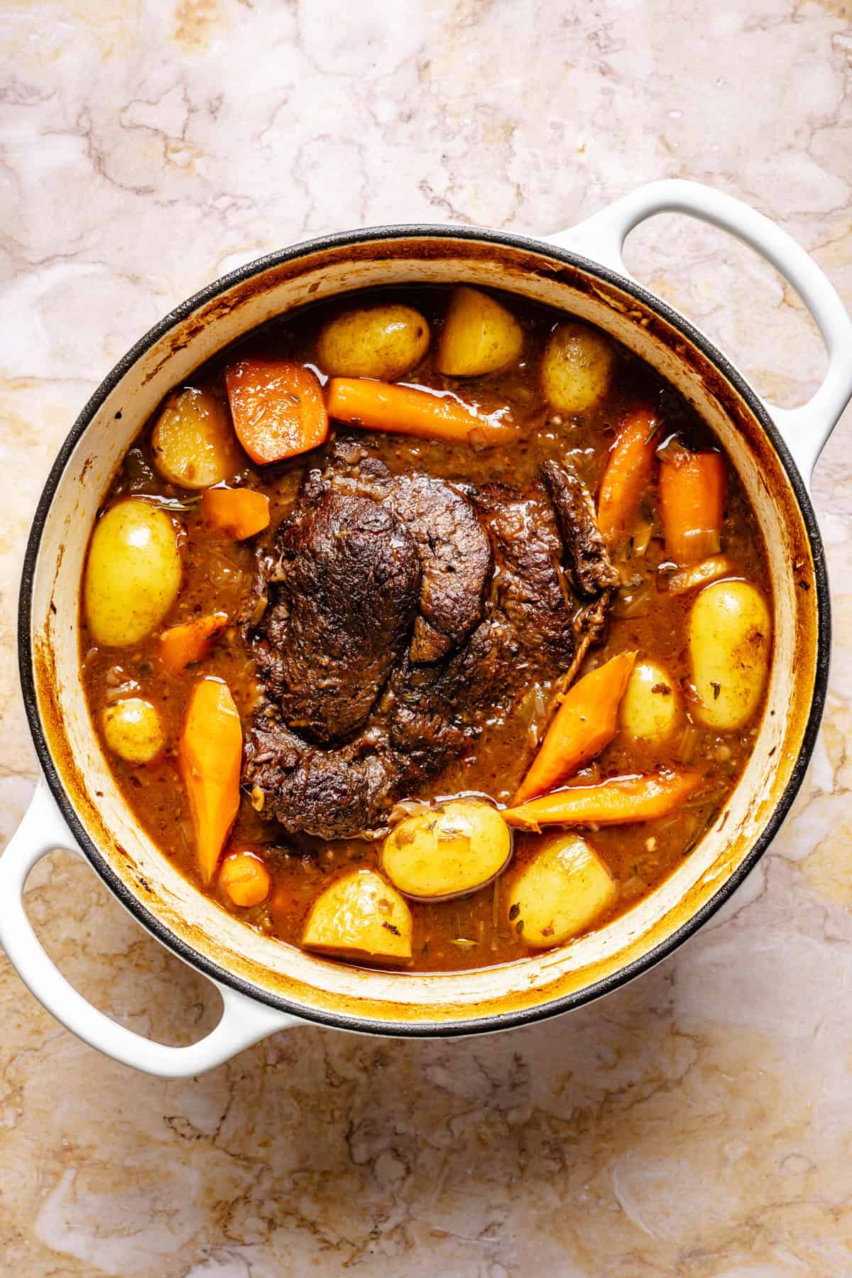 A white Dutch oven filled with pot roast, whole baby potatoes, carrots, and a rich brown broth, sits on a light marble surface.