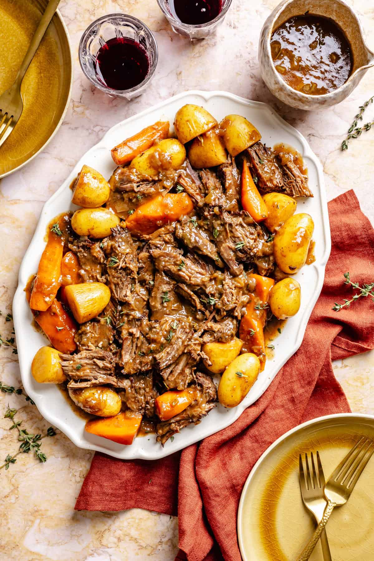 A serving platter of oven pot roast with chunks of beef, baby potatoes, and carrots in brown gravy, surrounded by plates, silverware, and glasses of red wine on a marble table with a red napkin.