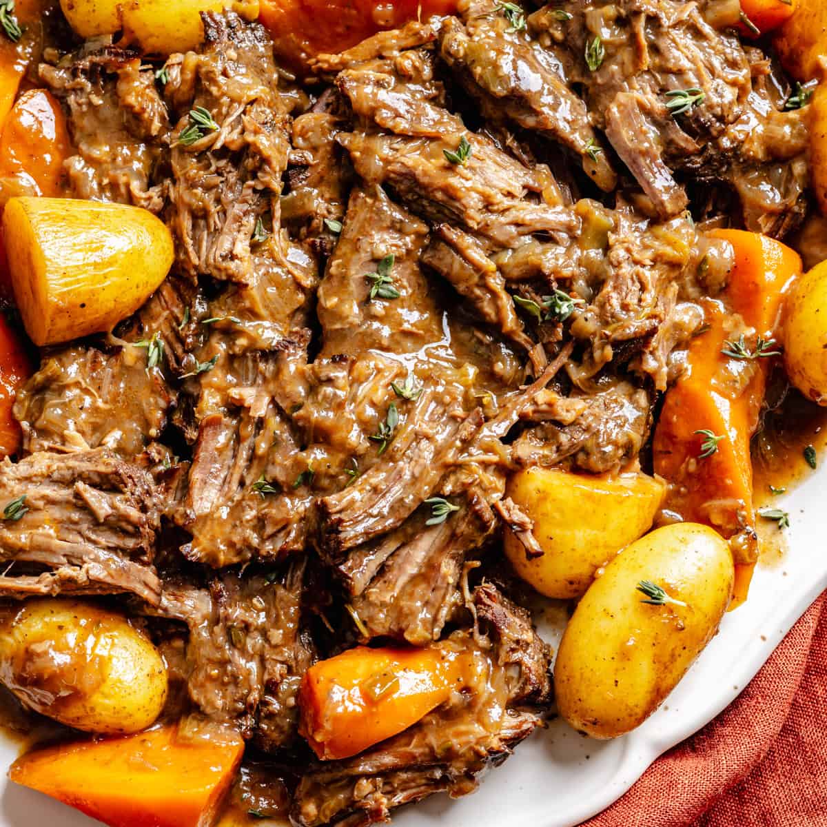 Oven Pot roast with shredded beef, gravy, baby potatoes, and carrots, garnished with fresh thyme on a white serving dish.