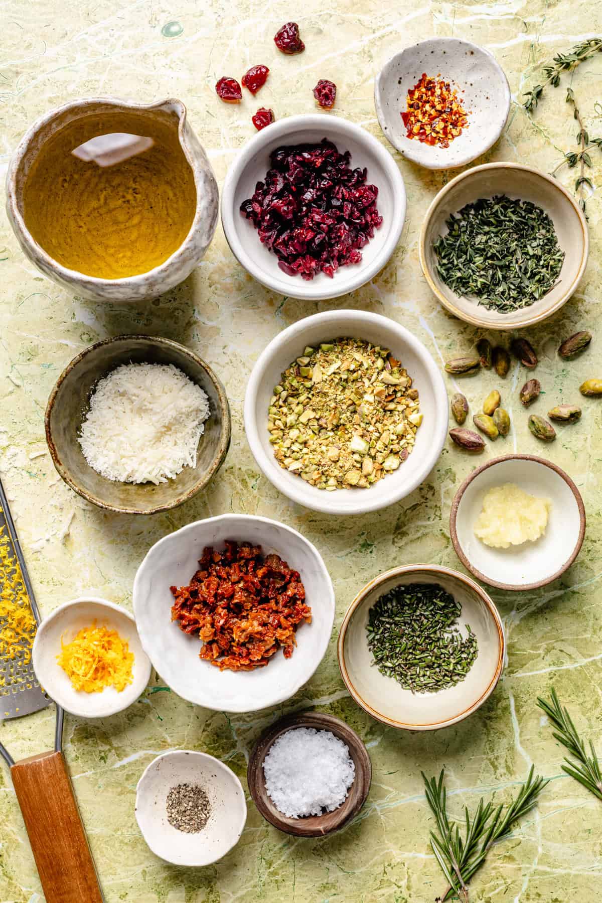 Small bowls arranged on a green marble surface contain ingredients including dried cranberries, chopped pistachios, sun-dried tomatoes, grated cheese, herbs, spices, salt, and olive oil.
