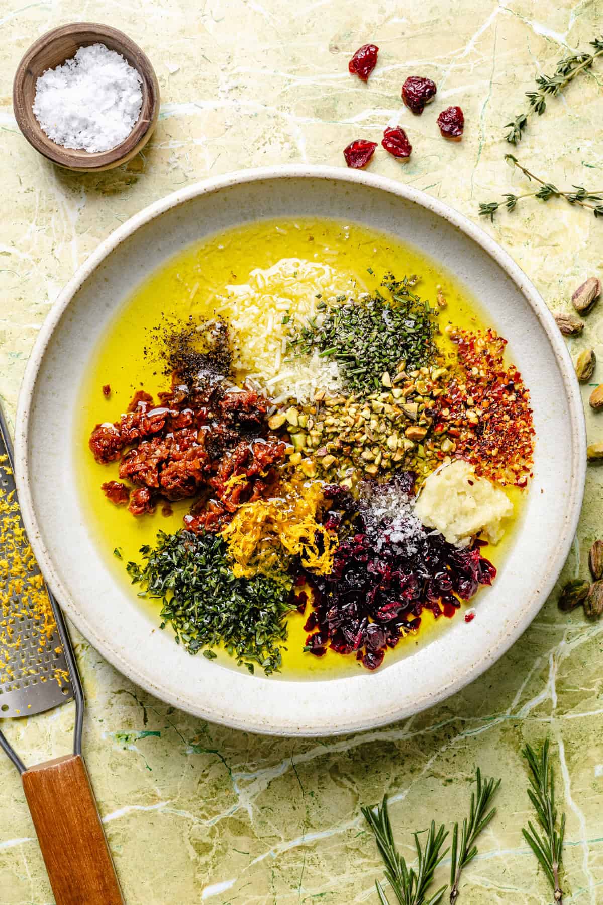 A shallow bowl filled with olive oil, topped with piles of chopped sun-dried tomatoes, fresh herbs, garlic, crushed nuts, red pepper flakes, citrus zest, and dried cranberries, with a small bowl of salt nearby for bread dipping oil.