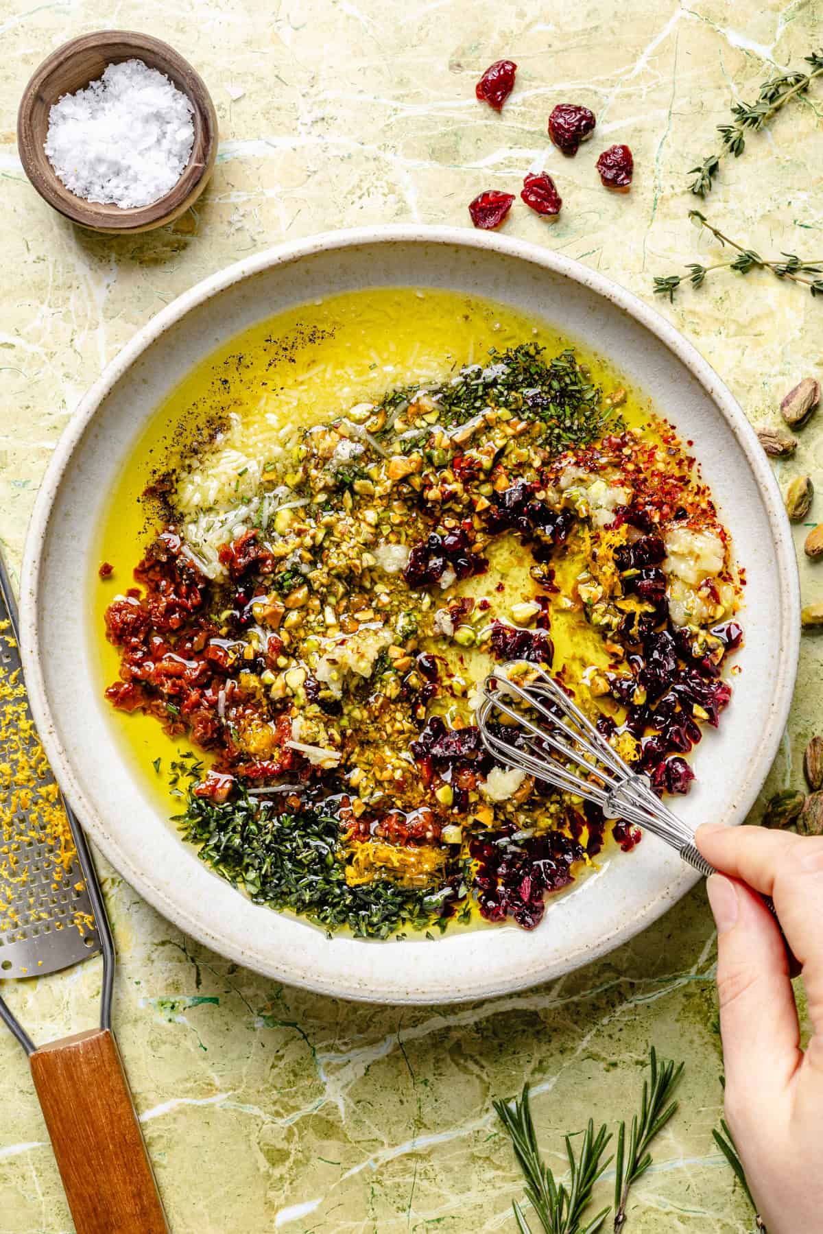 A hand uses a small whisk to stir a mixture of olive oil, chopped nuts, dried cranberries, herbs, and spices in a shallow bowl for bread dipping oil.