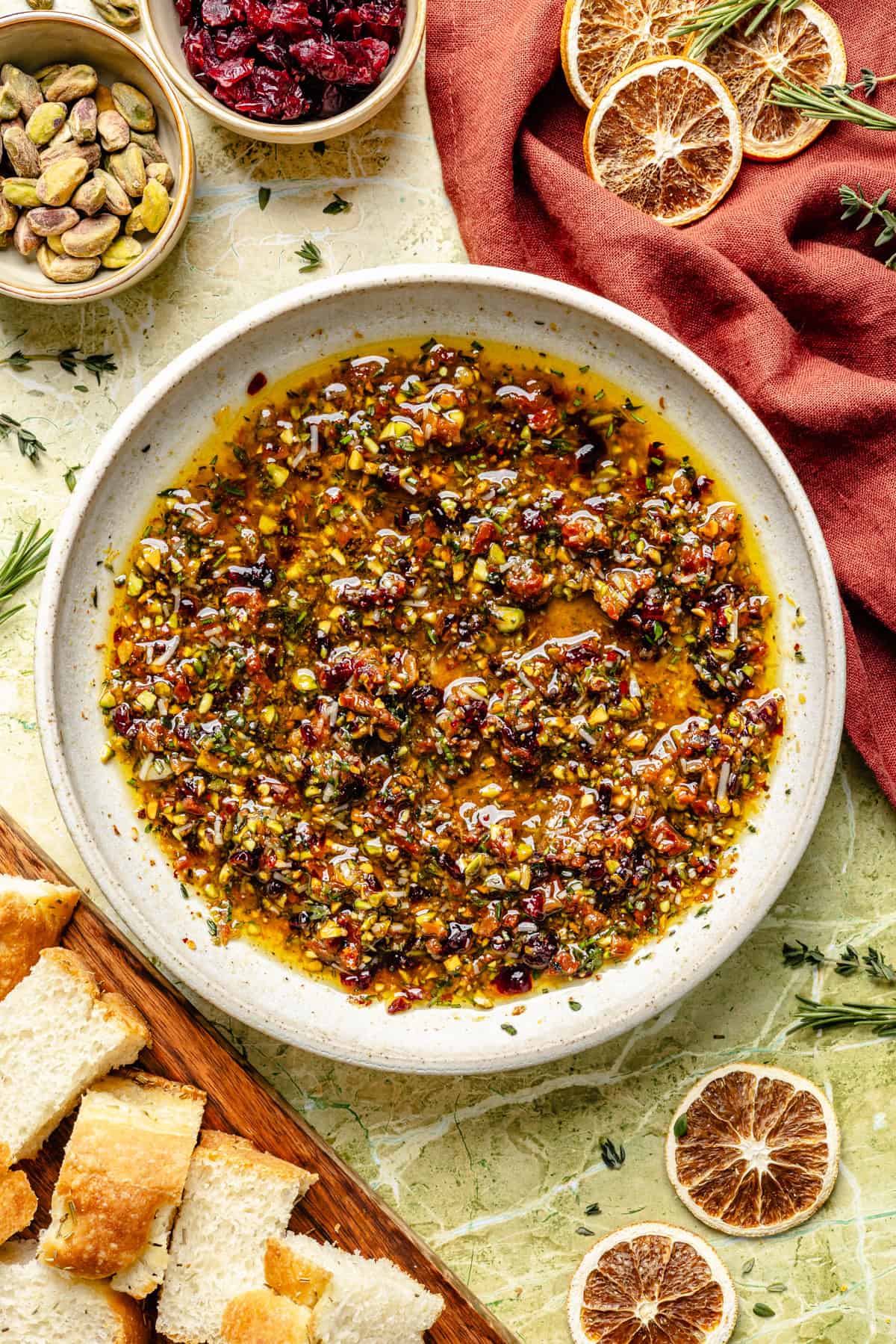 A bowl of dipping oil infused with chopped herbs, nuts, and dried fruit, served with slices of bread and garnished with dried orange slices.