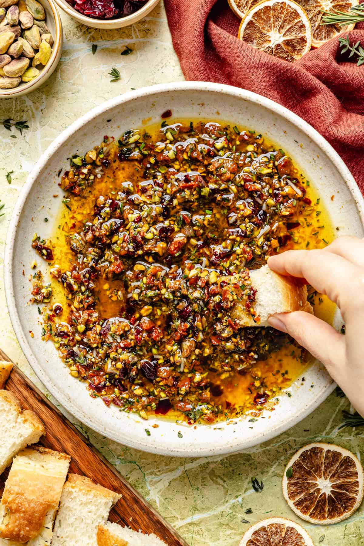 A hand dips a piece of bread into a bowl of herb and nut-infused olive oil, surrounded by bowls of pistachios, cranberries, and dried orange slices for bread dipping oil.