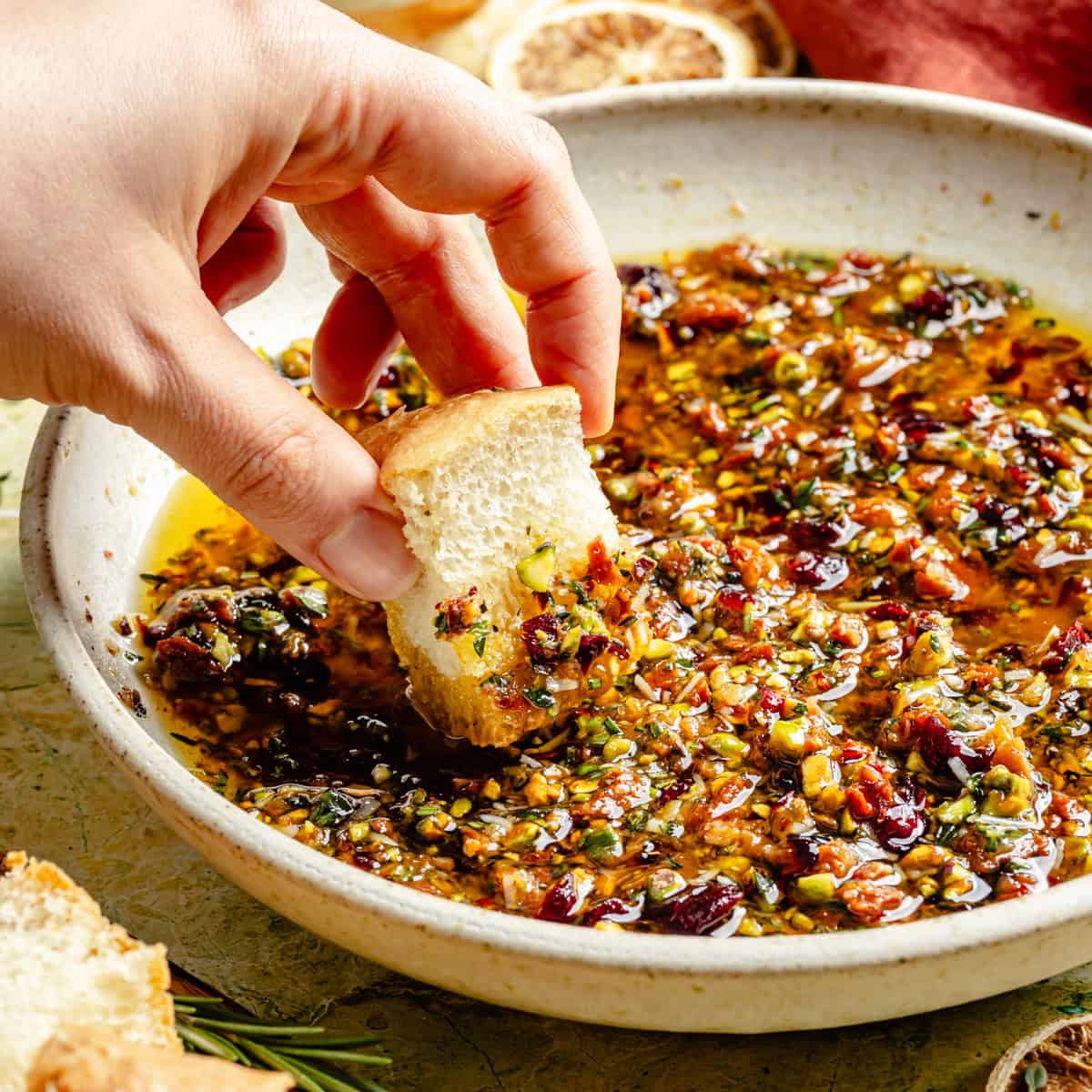 A hand dips a piece of crusty bread into a bowl of olive oil infused with chopped nuts, herbs, and sun-dried tomatoes for bread dipping oil.