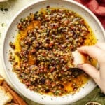 A hand dips a piece of crusty bread into a shallow bowl filled with an olive oil-based herb and sun-dried tomato bread dipping oil.