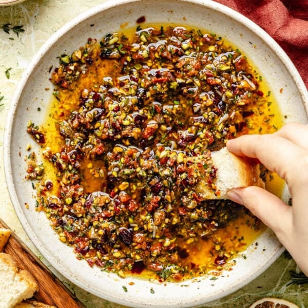 A hand dips a piece of crusty bread into a shallow bowl filled with an olive oil-based herb and sun-dried tomato bread dipping oil.