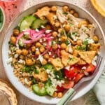 A greek chicken bowl topped with chickpeas, pita chips, cucumbers, cherry tomatoes, pickled red onions, and crumbled feta cheese.