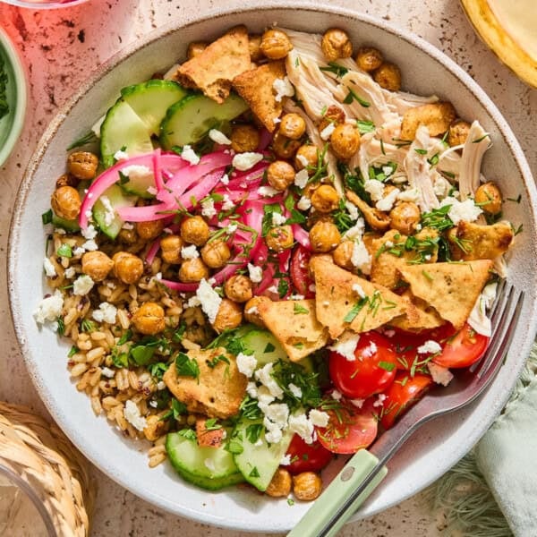 A greek chicken bowl topped with chickpeas, pita chips, cucumbers, cherry tomatoes, pickled red onions, and crumbled feta cheese.