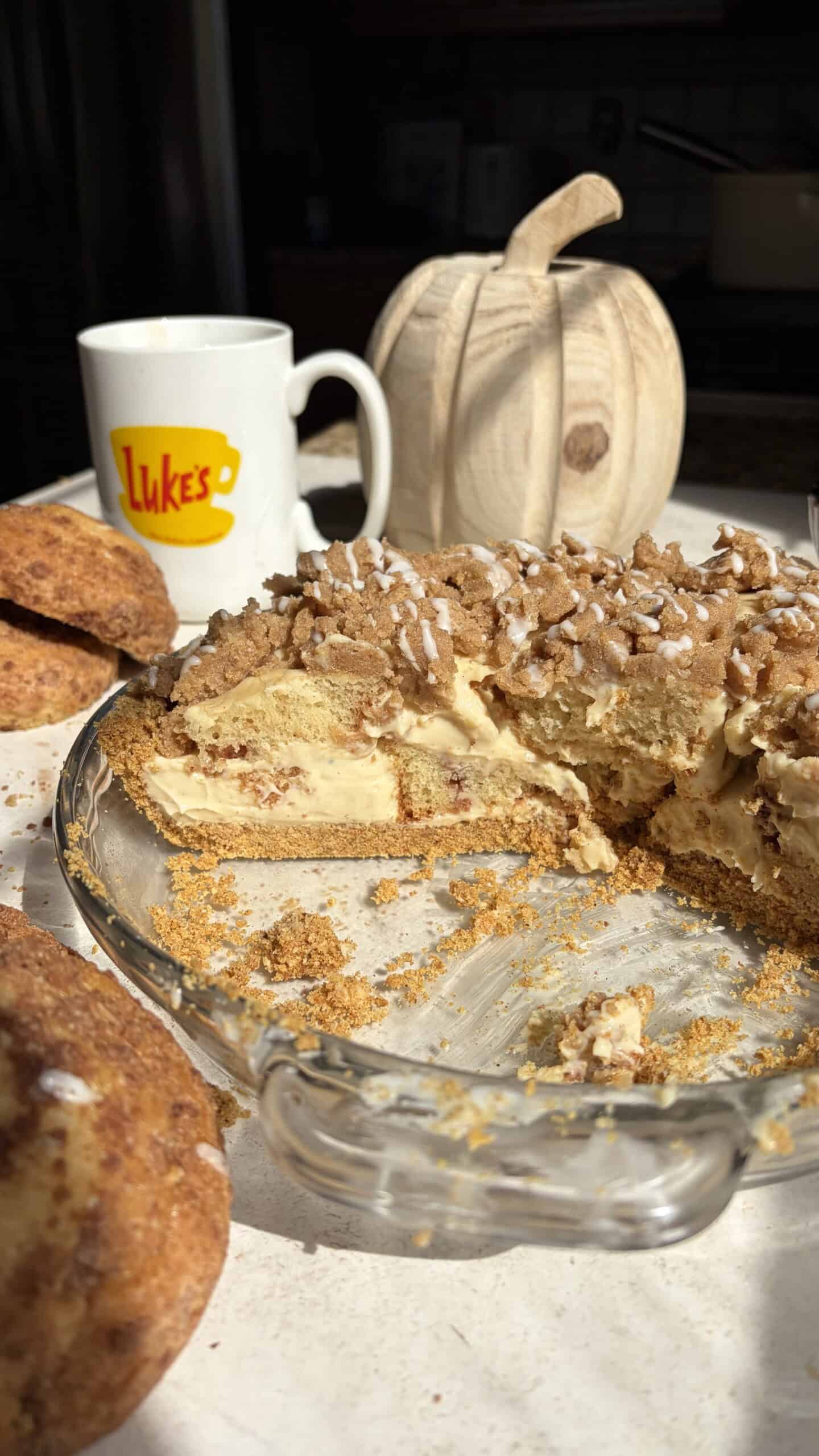 A pie with a crumbly streusel topping and creamy filling in a glass dish, set next to a mug labeled Lukes and a wooden pumpkin.