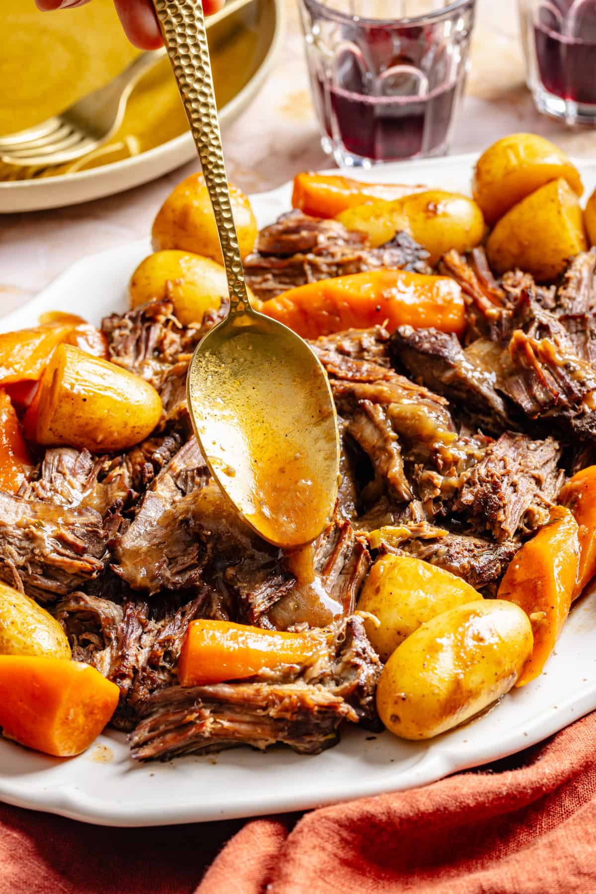 A gold spoon drizzling sauce over a platter of slow-cooked oven pot roast, potatoes, and carrots.