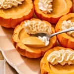 A spoon scoops creamy pumpkin posset from a small hollowed-out pumpkin topped with whipped cream.