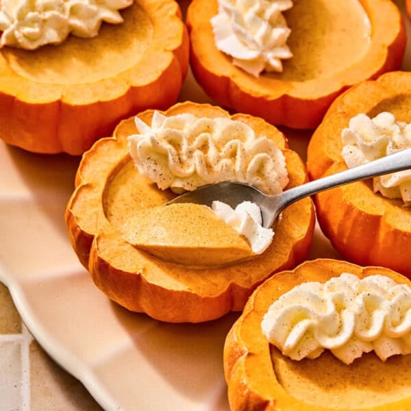 A spoon scoops creamy pumpkin posset from a small hollowed-out pumpkin topped with whipped cream.