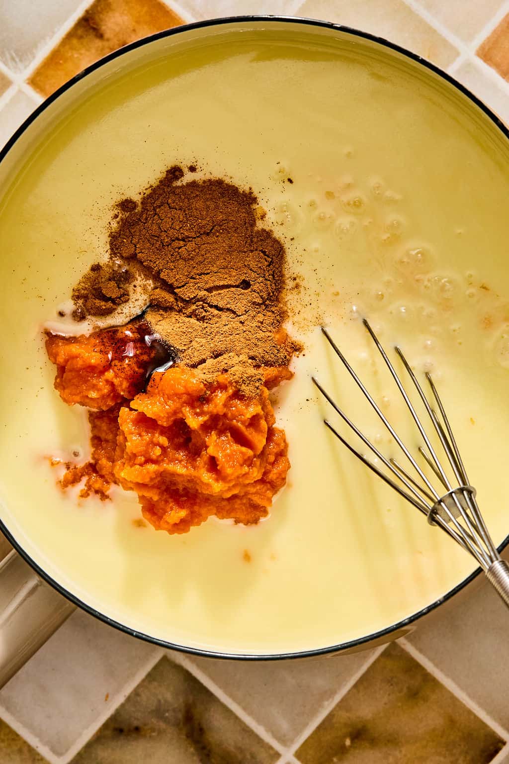 A whisk rests in a pot containing a creamy posset mixture, a scoop of orange pumpkin purée, and a dusting of brown spices.