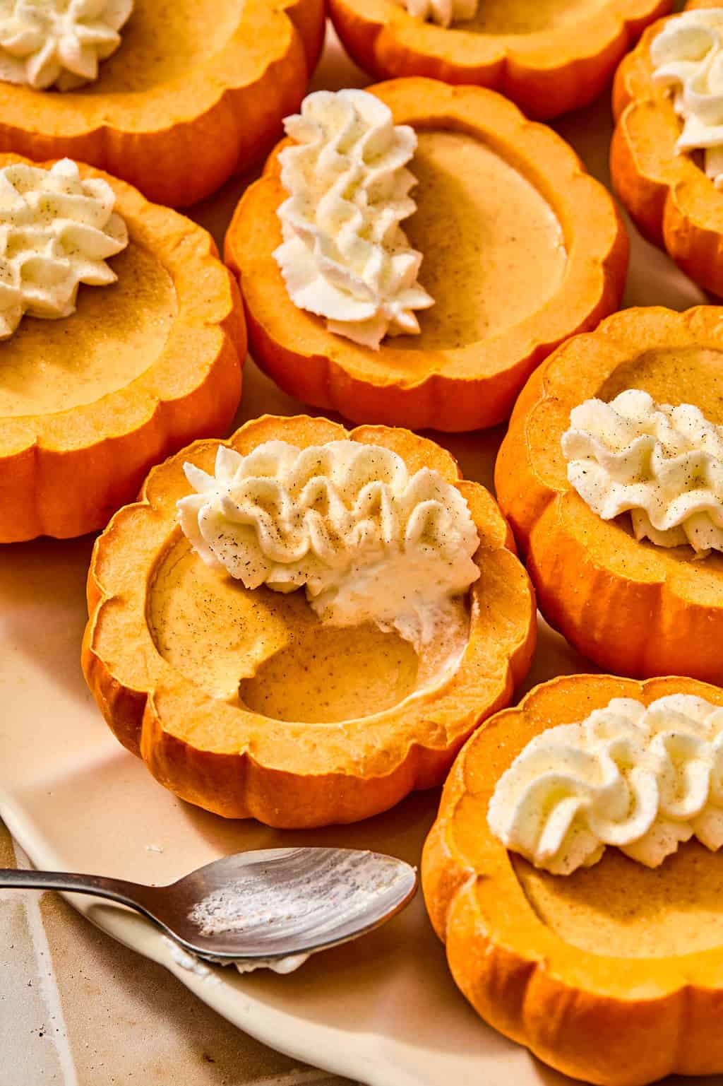 Several small pumpkins hollowed out and filled with pumpkin posset, topped with swirls of whipped cream, on a serving platter with a spoon.