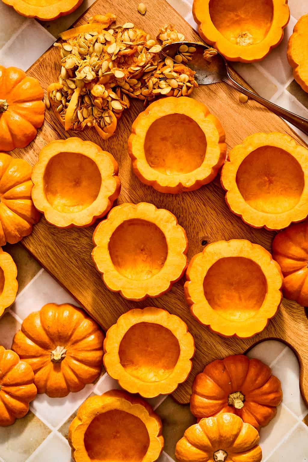 Several halved, hollowed-out mini pumpkins arranged on a wooden cutting board with a spoonful of pumpkin seeds nearby.