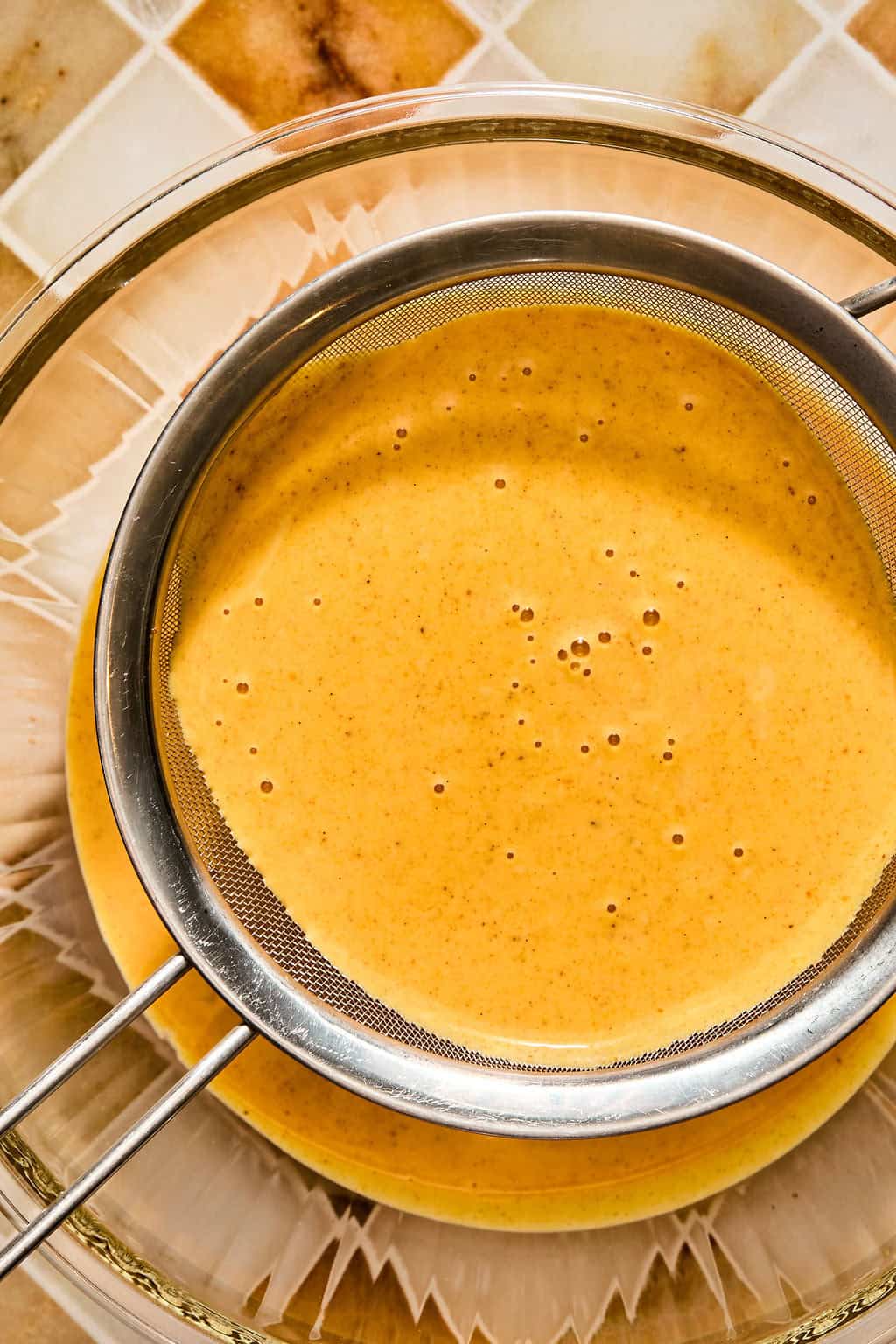 A metal mesh sieve sits over a glass bowl, straining the smooth pumpkin possset mixture.