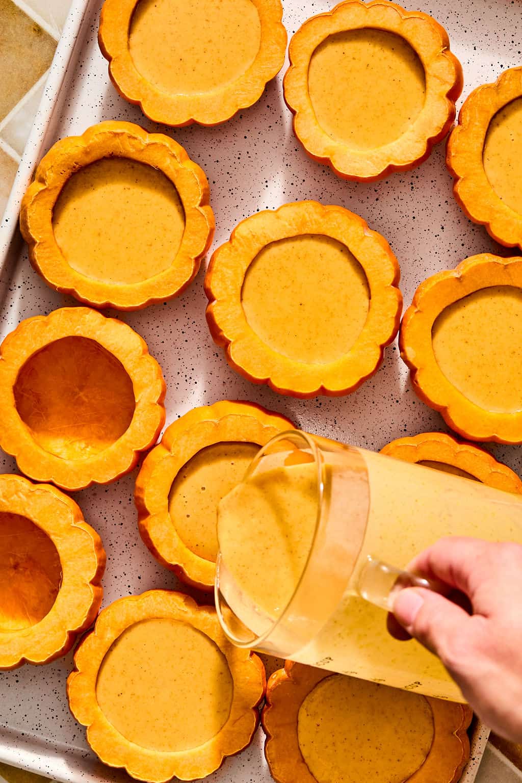 A hand pours pumpkin posset mixture from a glass pitcher into halved mini pumpkins arranged on a baking sheet.