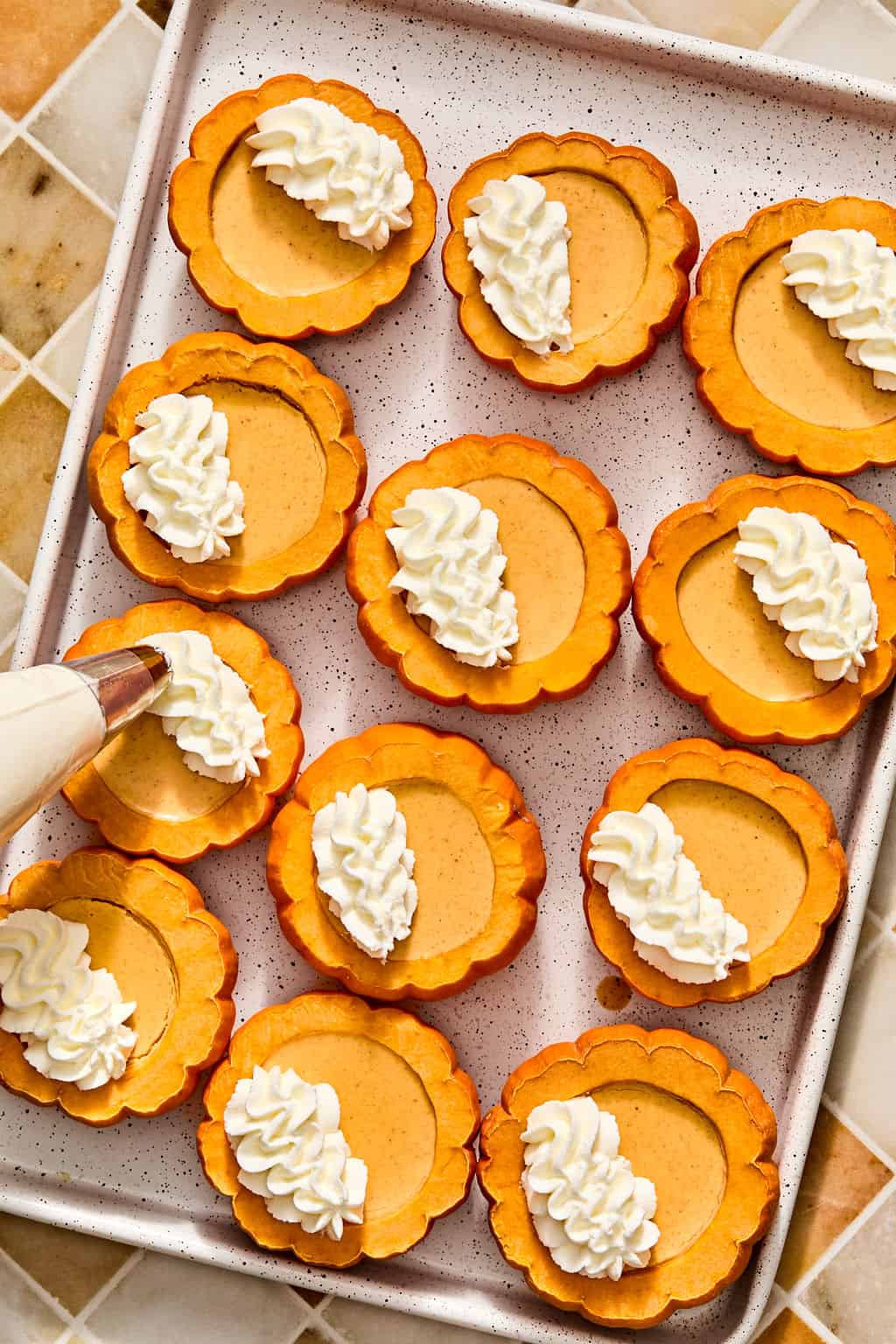 A piping bag dollops whipped cream onto pumpkin possets inside hollowed out mini pumpkins, arranged on a baking sheet.