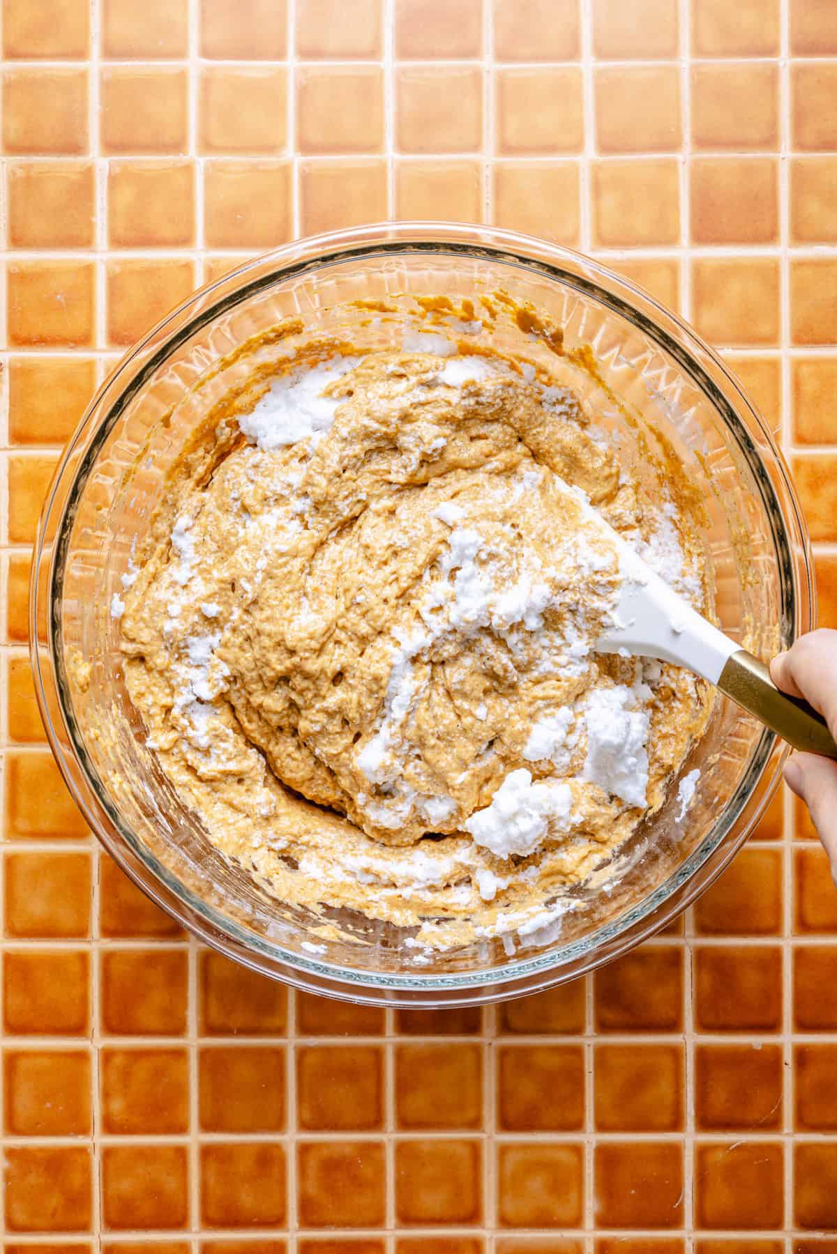 A hand folds whipped egg whites into pumpkin spiced pancake batter in a clear bowl on an orange tiled countertop.