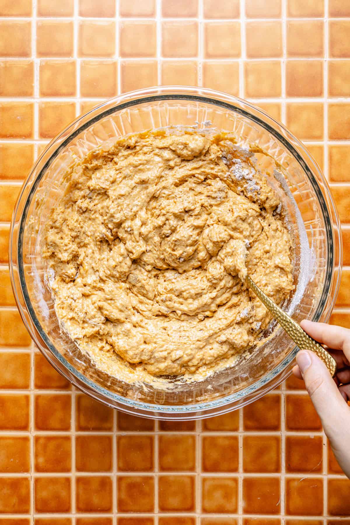 A hand stirs thick, pumpkin spiced pancake batter in a clear glass bowl on an orange tiled countertop.