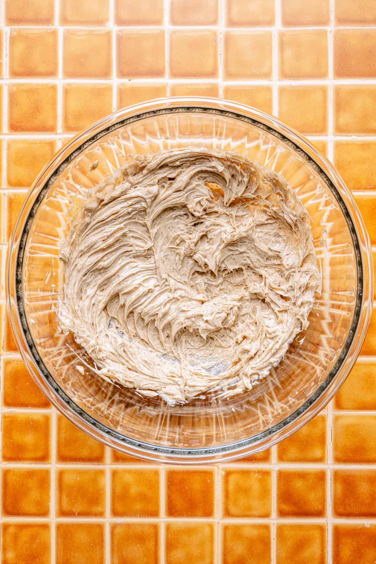 A glass bowl filled with light brown, whipped cinnamon butter sits on an orange tiled surface. The batter appears fluffy and well-mixed, with visible swirls from mixing.
