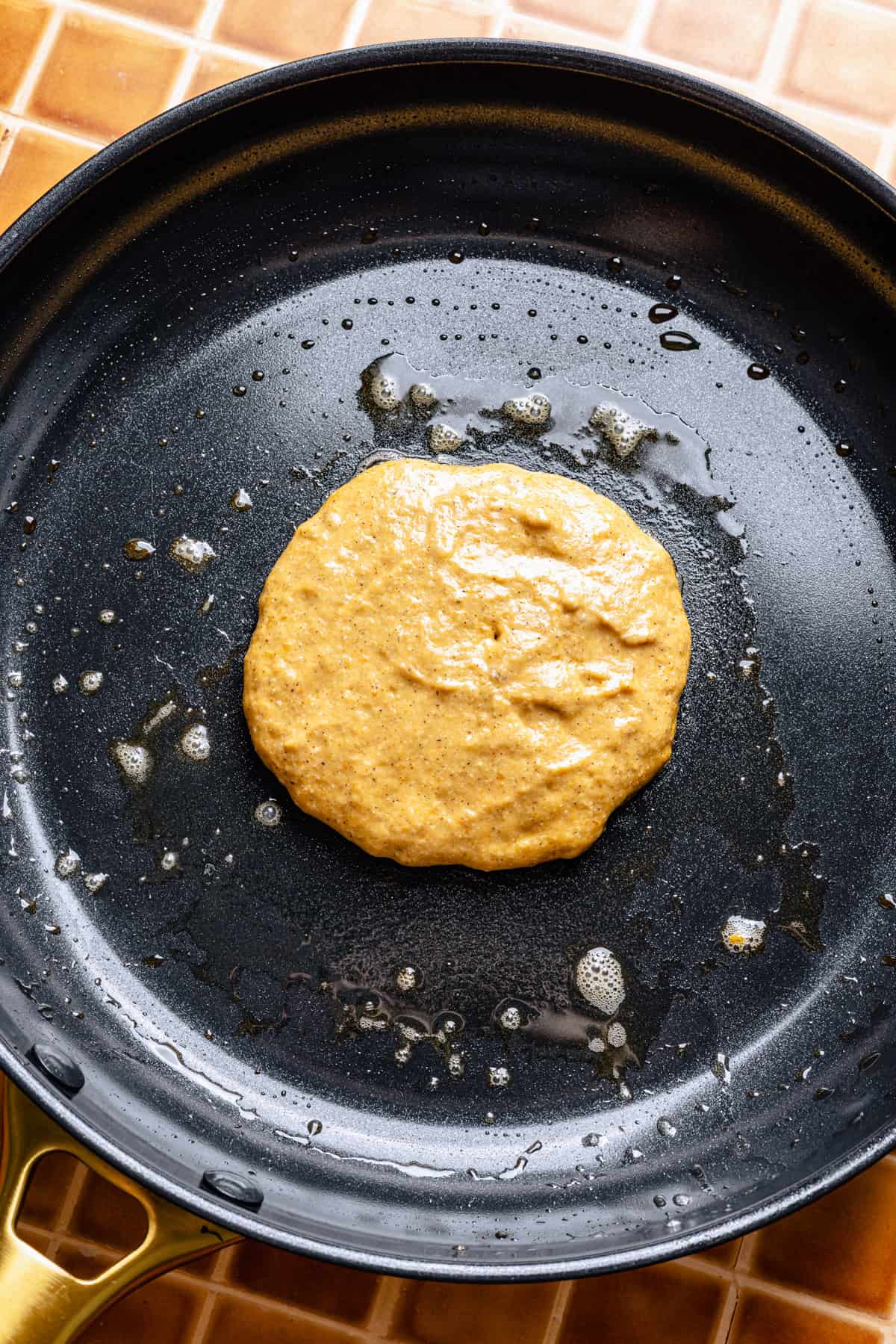 A dollop of pumpkin pancake batter cooks in a nonstick frying pan with melted butter on a brown tiled surface.