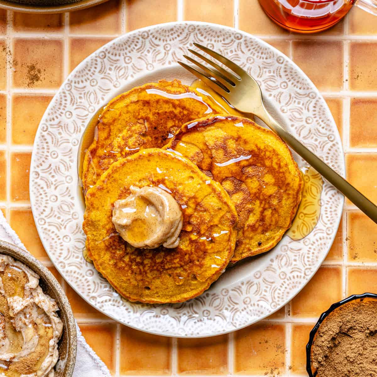 Three pancakes topped with a dollop of spiced butter and drizzled with syrup are served on a white patterned plate with a gold fork, set on an orange tiled background.