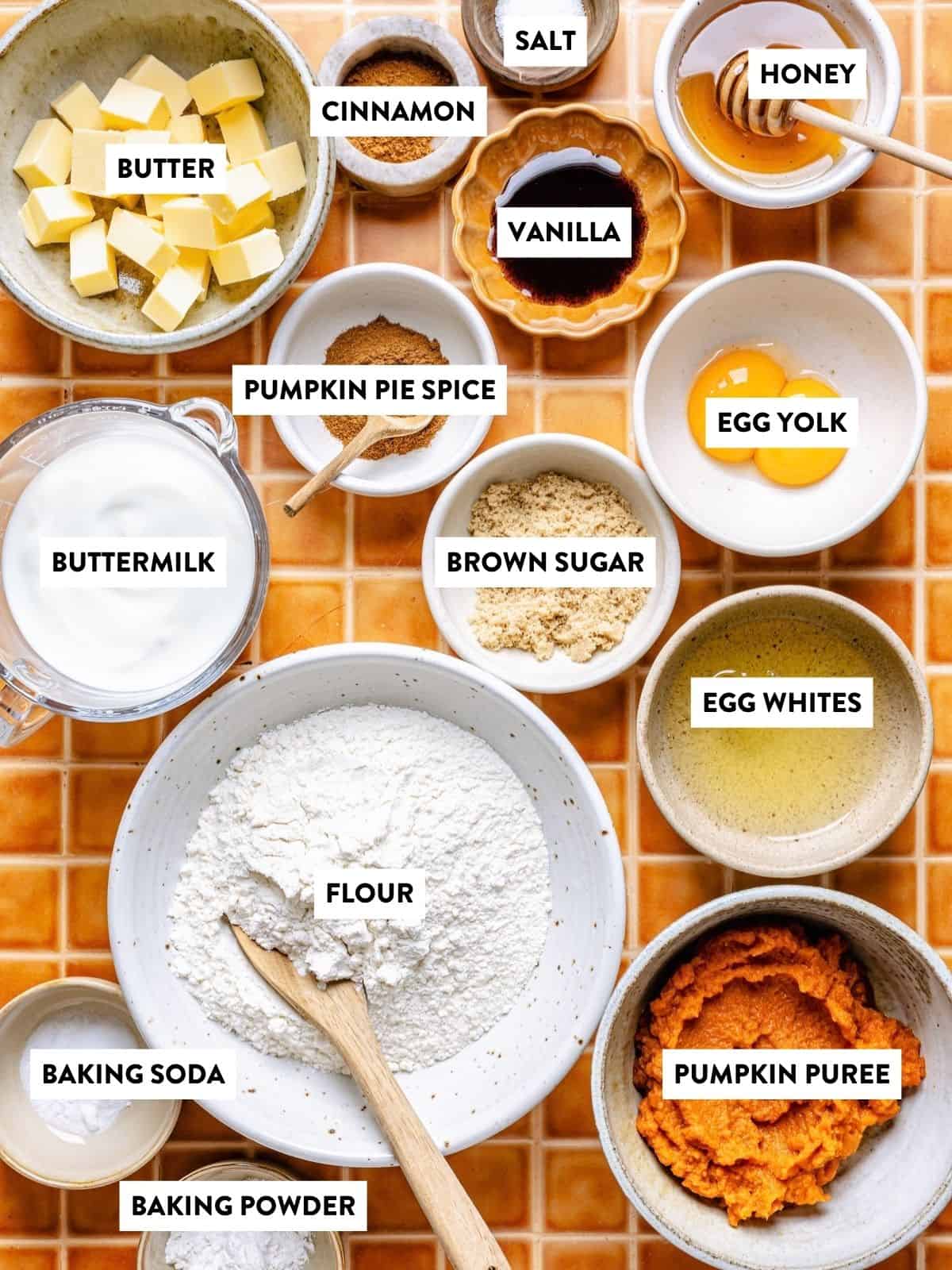 Various measured baking ingredients in bowls on an orange tile surface, including butter, cinnamon, salt, honey, vanilla, buttermilk, pumpkin pie spice, egg yolk, brown sugar, egg whites, flour, pumpkin puree, baking soda, and baking powder.