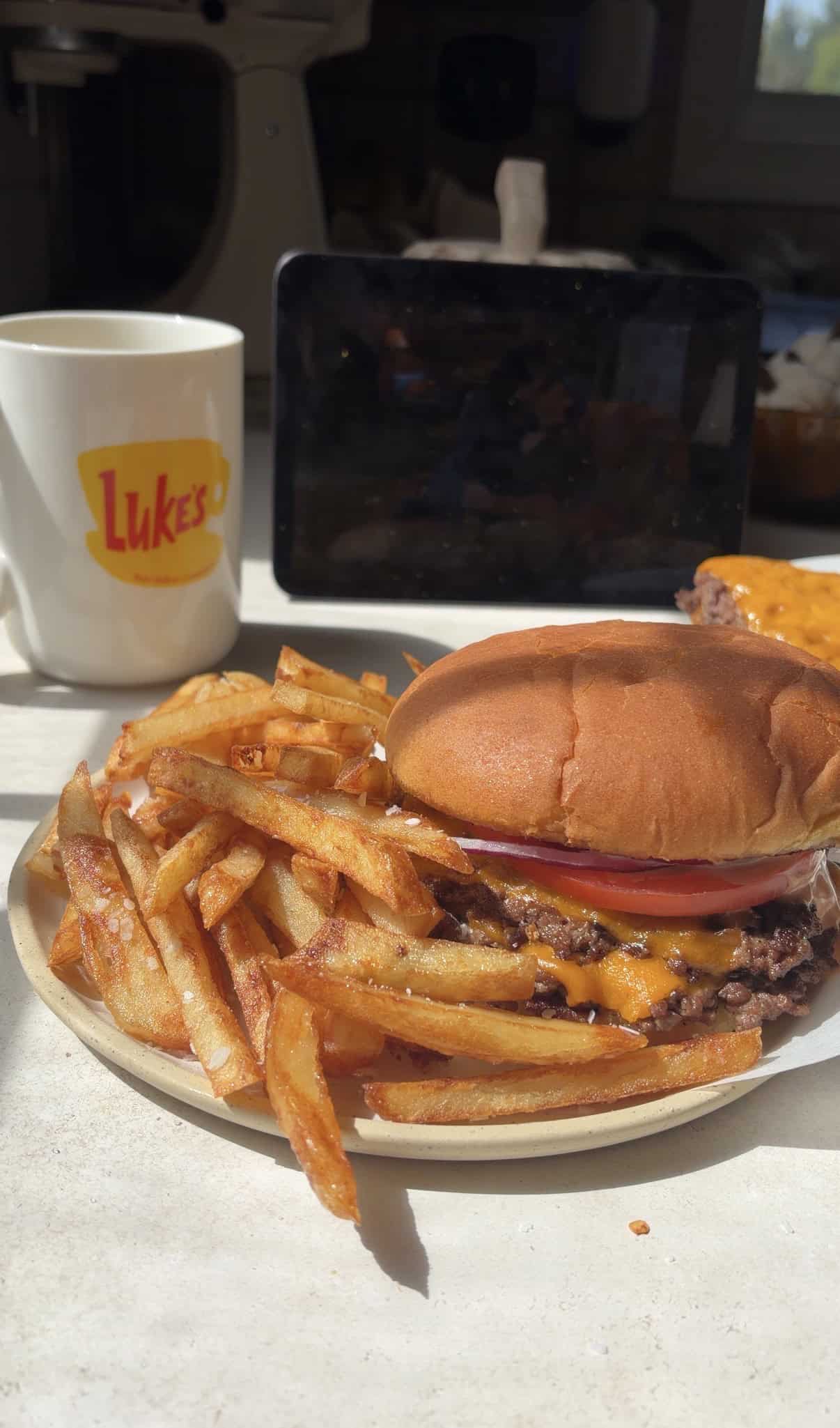 A cheeseburger and a pile of french fries on a plate, with a Lukes branded mug in the background.