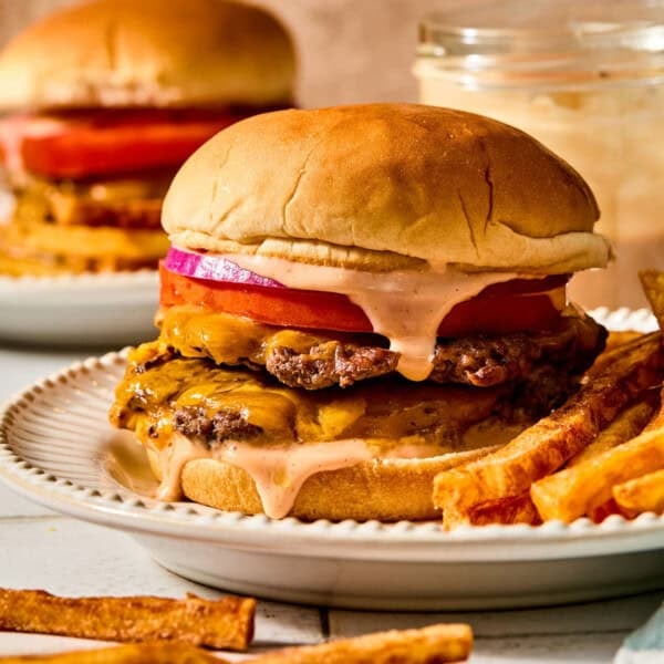 A double smash burger with red onion, tomato, and sauce on a ridged plate next to french fries, with another burger blurred in the background.