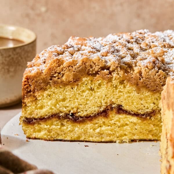 A slice of sour cream coffee cake with a crumb topping dusted with powdered sugar on a plate next to a cup of coffee.